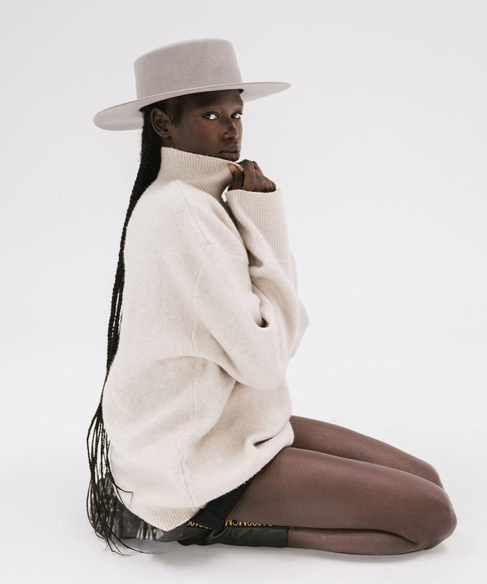 Woman with long hair wearing a light grey boater-style hat and a cream sweatshirt, sitting against a plain background #color_light grey