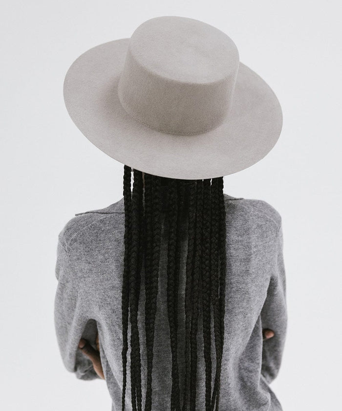 Woman with long hair wearing a light grey boater-style hat and a grey shirt, facing behind against a plain background #color_light grey