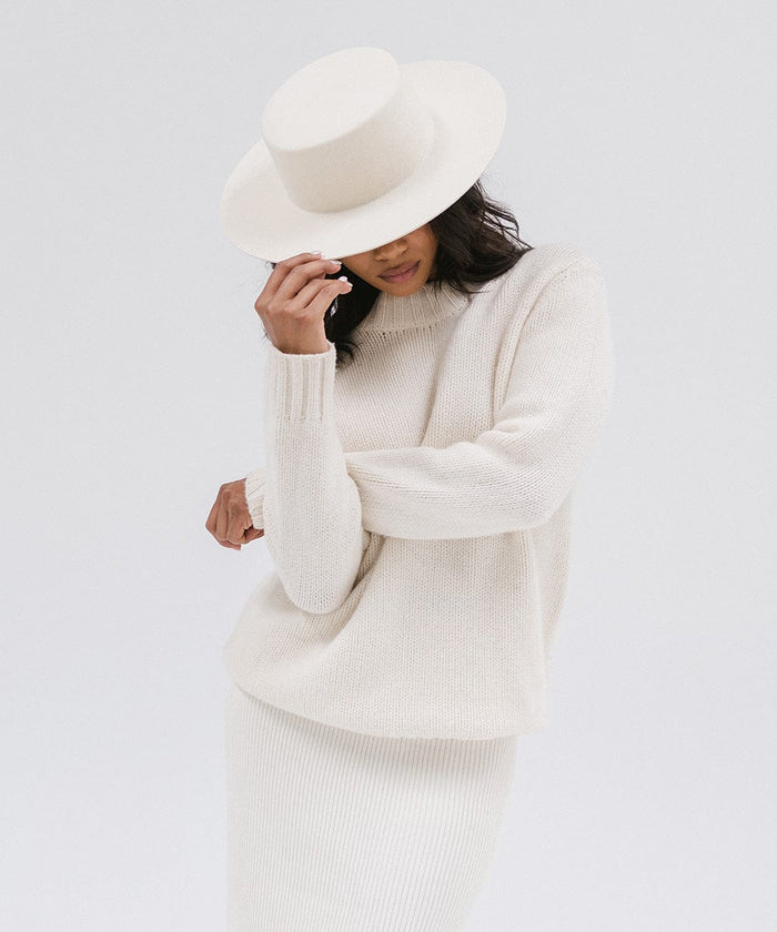 Woman wearing an off white boater-style hat, a white sweatshirt and white skirt against a plain background #color_off white