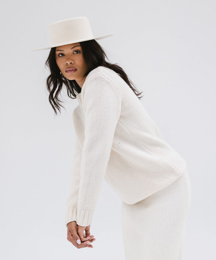 Woman wearing an off white boater-style hat, a white sweatshirt and white skirt, posing against a plain background #color_off white