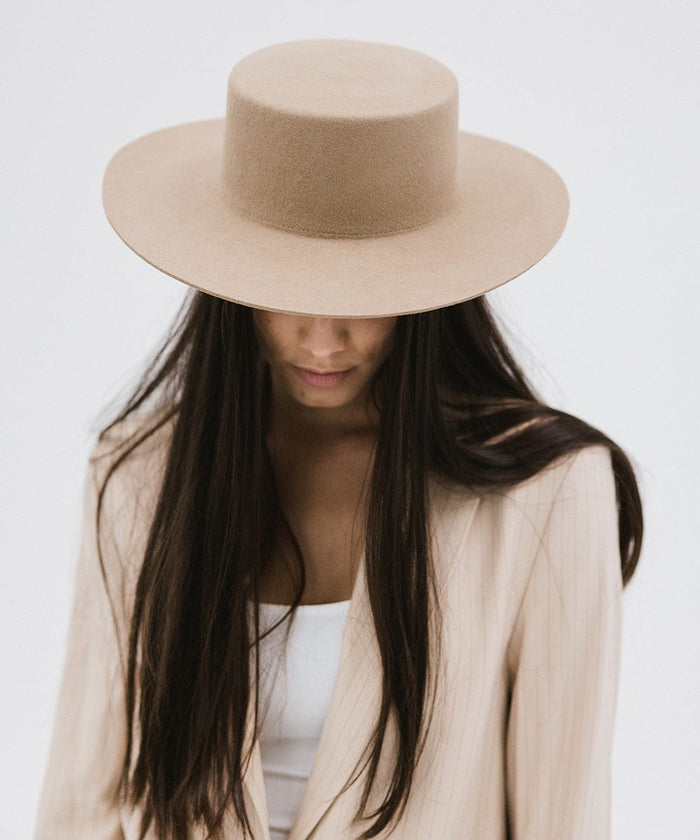Woman with long hair wearing a tan boater-style hat and a cream blazer, looking down against a plain background #color_tan