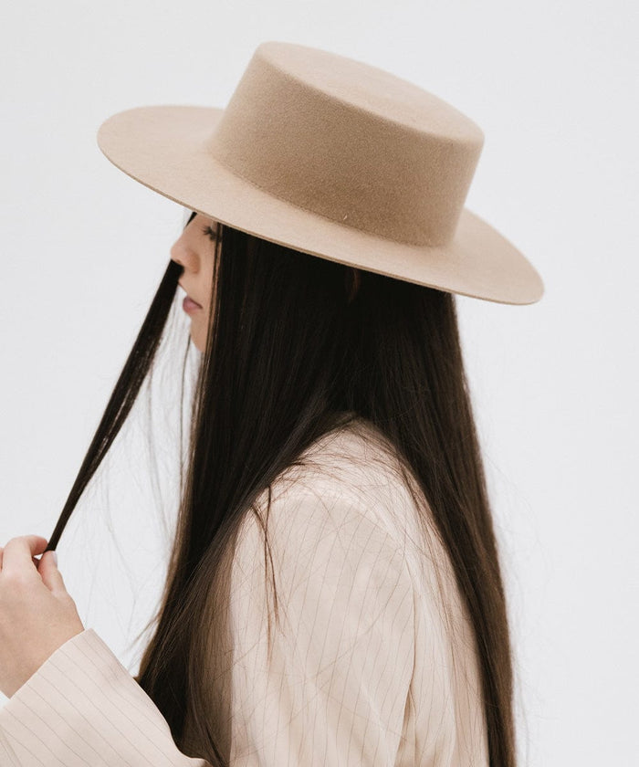 Woman with long hair wearing a tan boater-style hat and a cream blazer, facing sideways against a plain background #color_tan