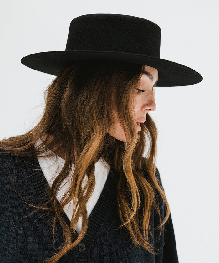 Woman wearing a black boater-style hat and a dark blue shirt, looking sideways against a plain background #color_black