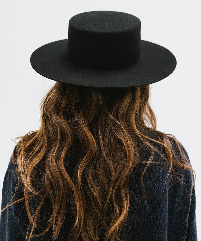 Woman with long hair wearing a black boater-style hat and a dark blue shirt, facing behind against a plain background #color_black