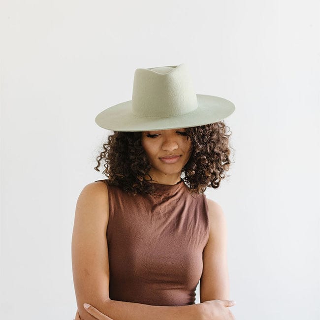 Woman wearing a light green fedora hat and a brown top, looking down against a plain background #color_light green