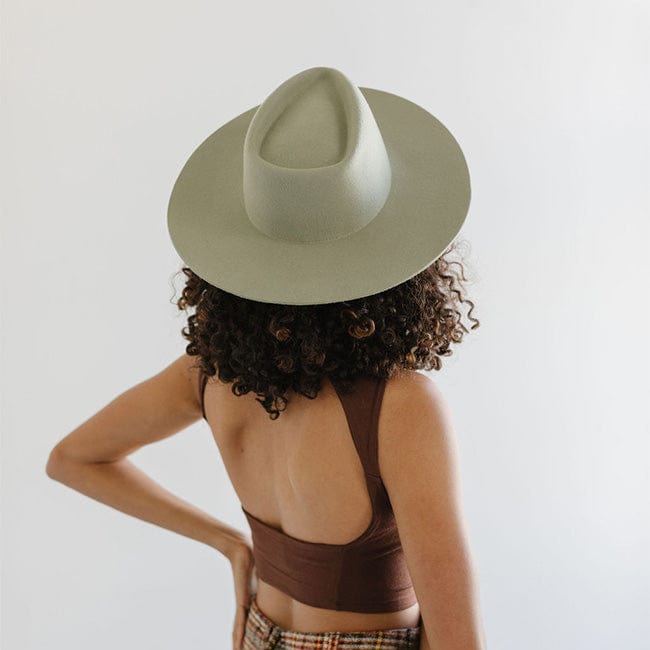 Woman wearing a light green fedora hat and a brown top, facing behind against a plain background #color_light green