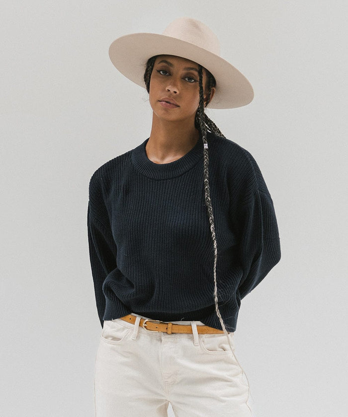 Woman wearing a cream wide brim felt hat with a triangle crown, a dark blue sweatshirt, brown belt, and white pants against a white background #color_cream