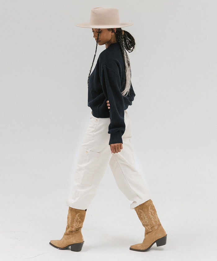 Woman wearing a cream wide brim felt hat with a triangle crown, a dark blue sweatshirt, brown belt, white pants, and brown boots, posing against a white background #color_cream