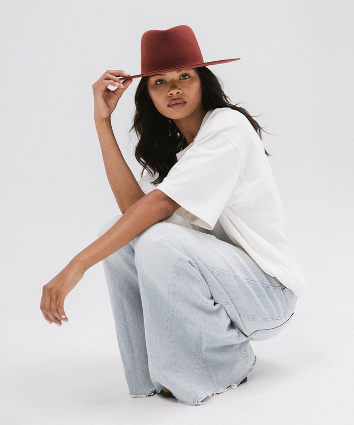 Woman wearing a rusty red wide brim felt hat with a triangle crown, a white t-shirt, and jeans, sitting against a white background #color_rusty red