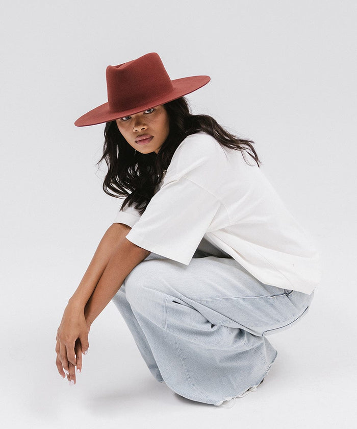 Woman wearing a rusty red wide brim felt hat with a triangle crown, a white t-shirt, and jeans, sitting against a white background #color_rusty red