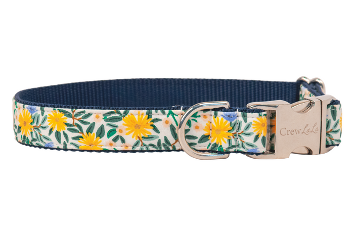 Dog collar with a light background and floral print featuring bright yellow daisies, pale colored blossoms, and leaves, finished with a metal buckle engraved with “Crewlala” and a matching D-ring.
