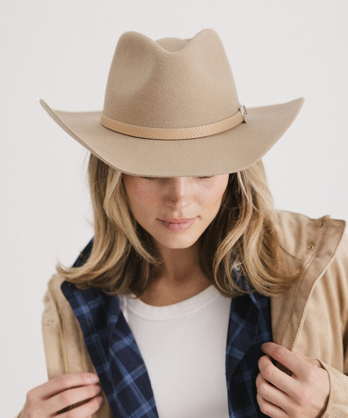 Woman wearing a tan cowboy hat with a hat band and a white top with a beige jacket against a plain background #color_tan
