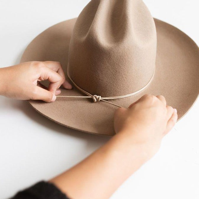 Person setting a tan double strand rope hat band on a beige hat on a plain background #color_tan