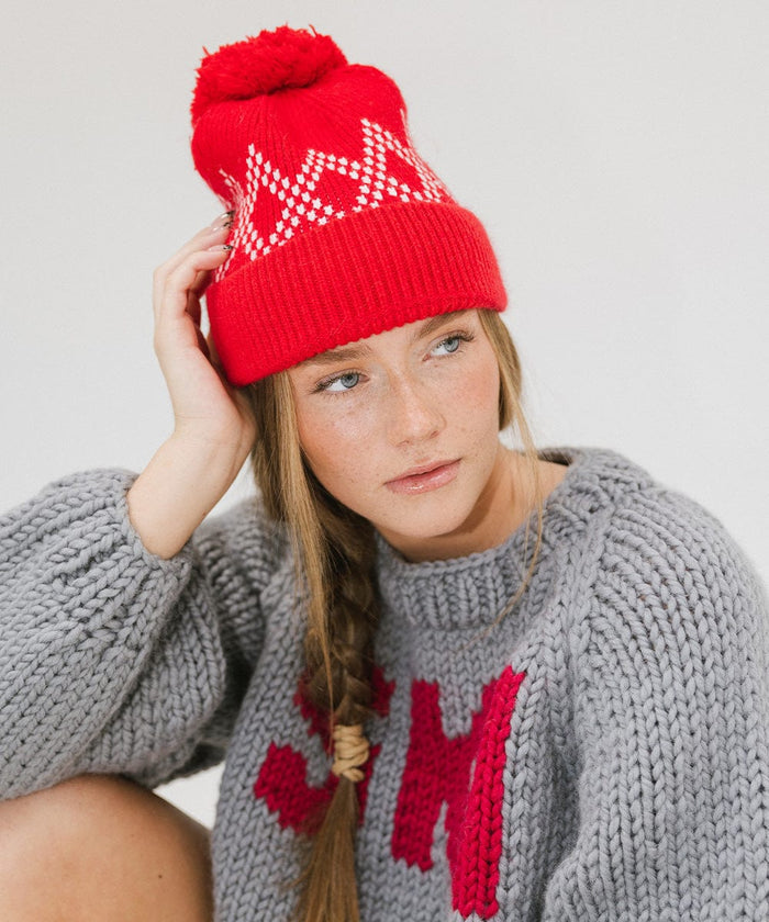 Woman wearing a ruby red colored knit beanie with a pom-pom and a grey sweater, looking sideways against a plain background #color_ruby red