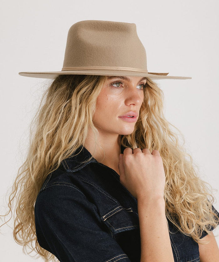 Woman with long blonde hair wearing a tan fedora hat and a denim shirt, looking sideways against a plain background  #color_tan