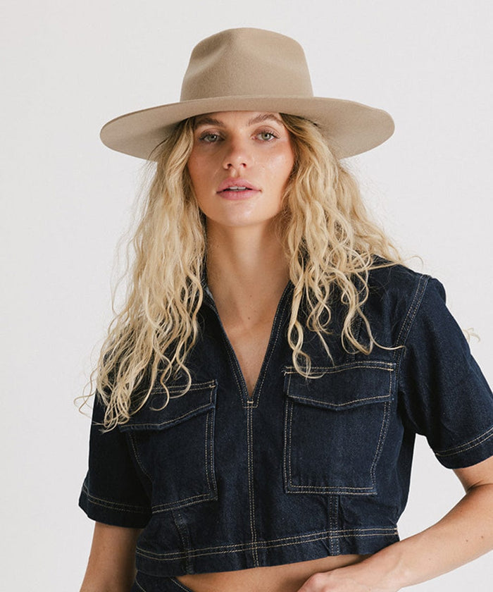 Woman with long blonde hair wearing a tan fedora hat and a denim top against a plain background  #color_tan