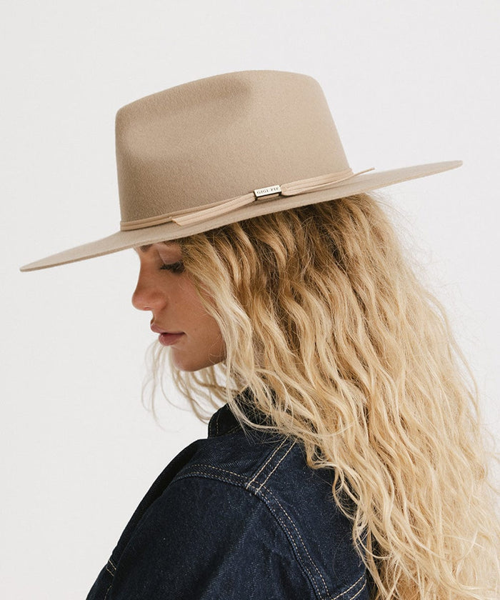 Woman with blonde hair wearing a tan fedora hat with a hat band and a denim shirt, facing sideways against a plain background  #color_tan