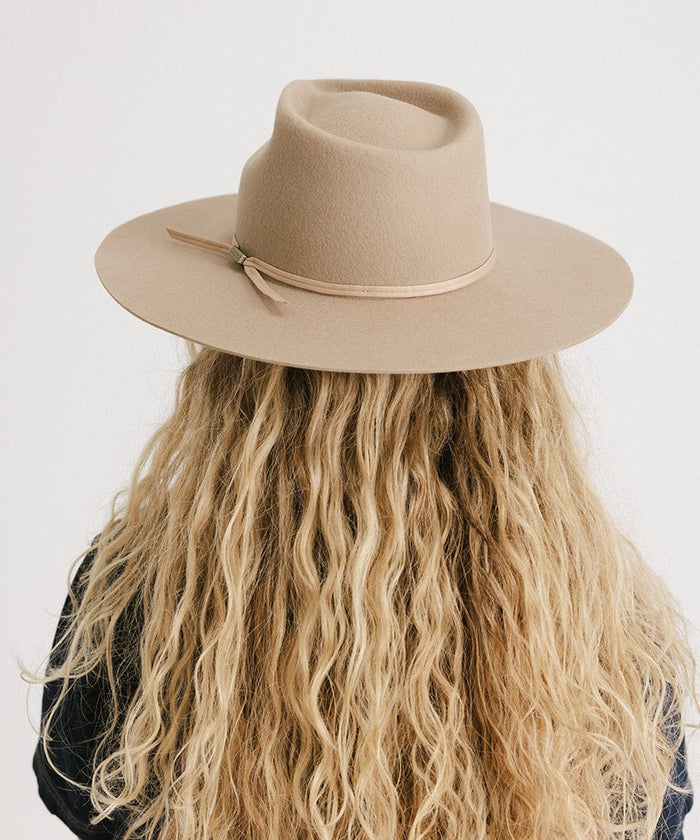 Woman with long blonde hair wearing a tan fedora hat with a hat band facing behind against a plain background  #color_tan