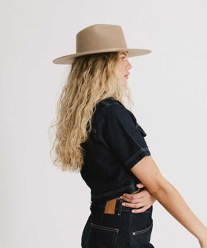 Woman with long blonde hair wearing a tan fedora hat, a denim shirt and jeans, facing sideways against a plain background  #color_tan