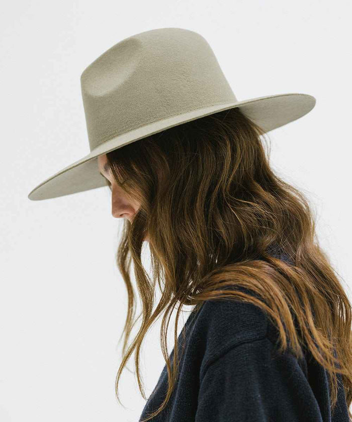 Woman wearing a sage colored wide brim fedora hat and a blue sweater, facing sideways against a plain background #color_sage