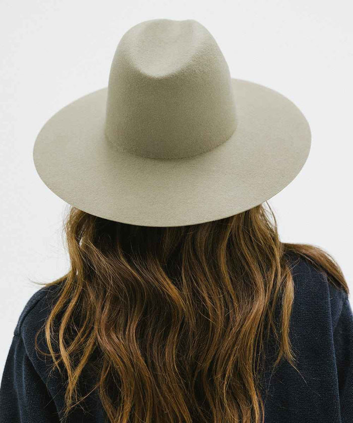 Woman wearing a sage colored wide brim fedora hat and a blue sweater, facing behind against a plain background #color_sage