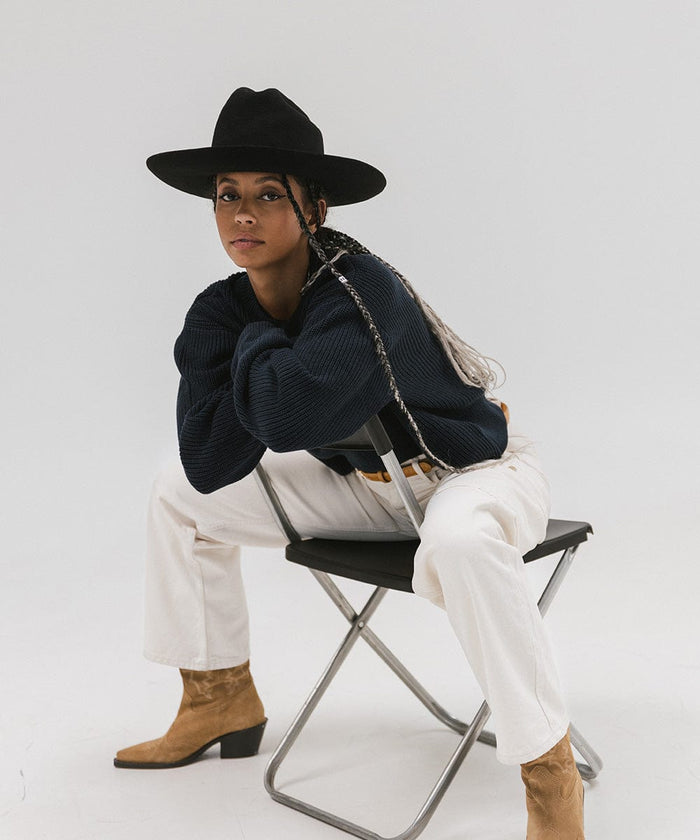 Woman wearing a black Western hat with a classic cattleman crown, a dark blue sweatshirt, white pants, and brown boots, sitting on a chair  against a plain background  #color_black