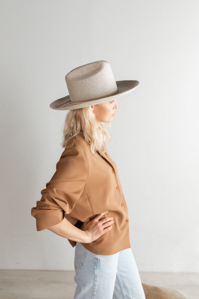 Woman wearing a mix grey colored wide brim felt hat with a hat band, a brown shirt and light blue jeans, facing sideways against a plain background #color_mix grey
