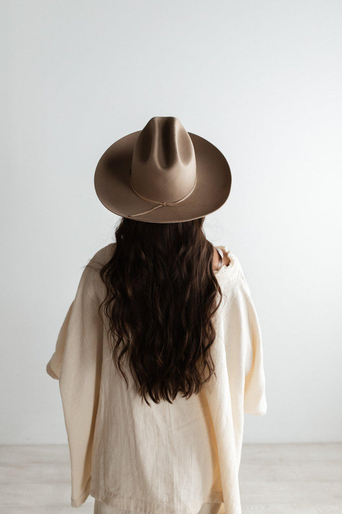 Woman wearing a tan cowboy hat with a hat band and a white dress, facing behind against a plain background #color_tan