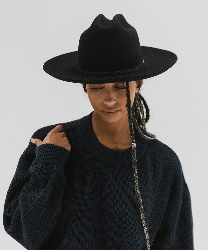 Woman wearing a black Western hat with a classic cattleman crown and a black hat band, and a dark blue sweatshirt, looking down against a plain background  #color_black