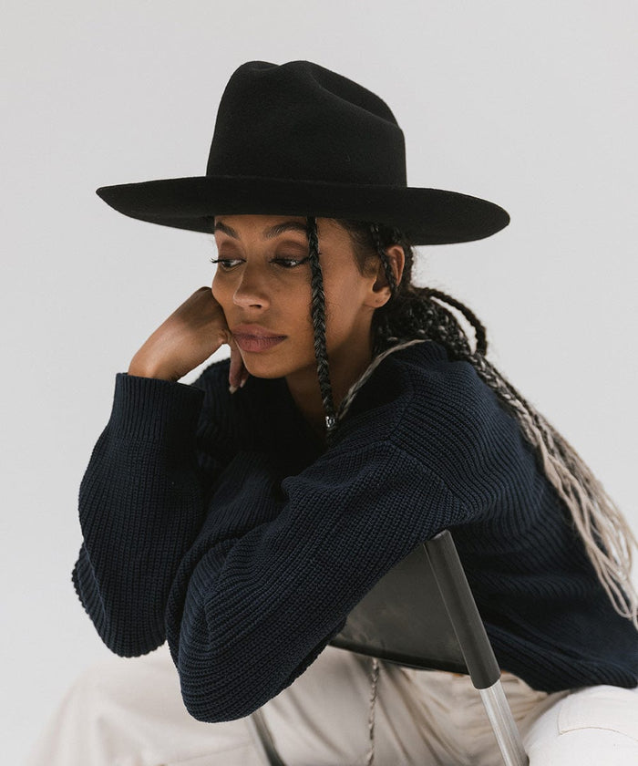 Woman wearing a black Western hat with a classic cattleman crown, and a dark blue sweatshirt sitting on a chair  against a plain background  #color_black