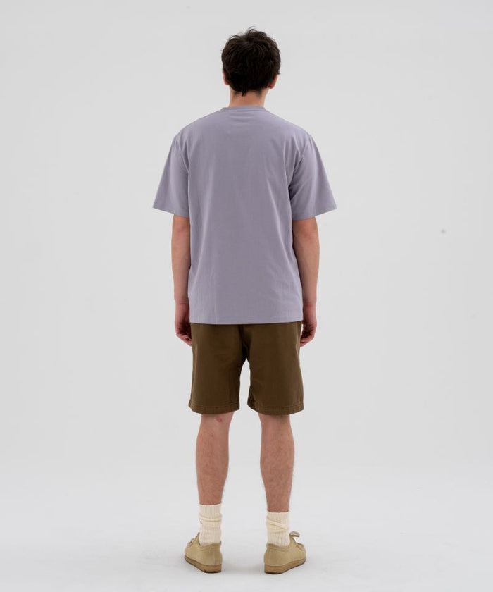 Back view of a model wearing a lavender Gramicci Half Dome Tee and brown shorts on a light-tone background.