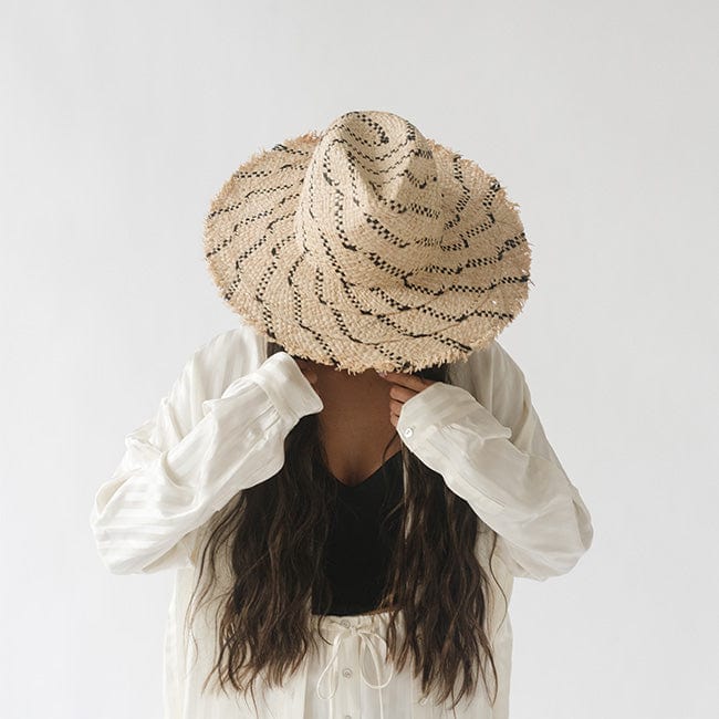 Woman wearing a natural colored straw hat with black patterns, a black top with a white shirt and white pants against a plain background #color_natural/black