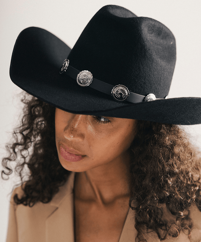 Woman wearing a black cowboy hat with decorative band and conchos on a neutral background #color_black