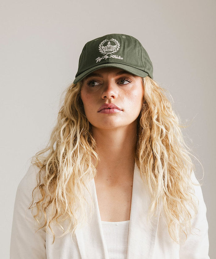 Woman wearing a dark green ball cap with a white logo and text and a white blazer against a plain background  #color_dark green