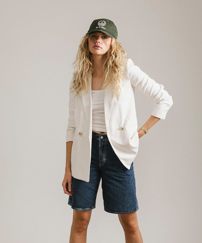 Woman wearing a dark green ball cap with a white logo and text, a white top with a white blazer and denim shorts against a plain background  #color_dark green