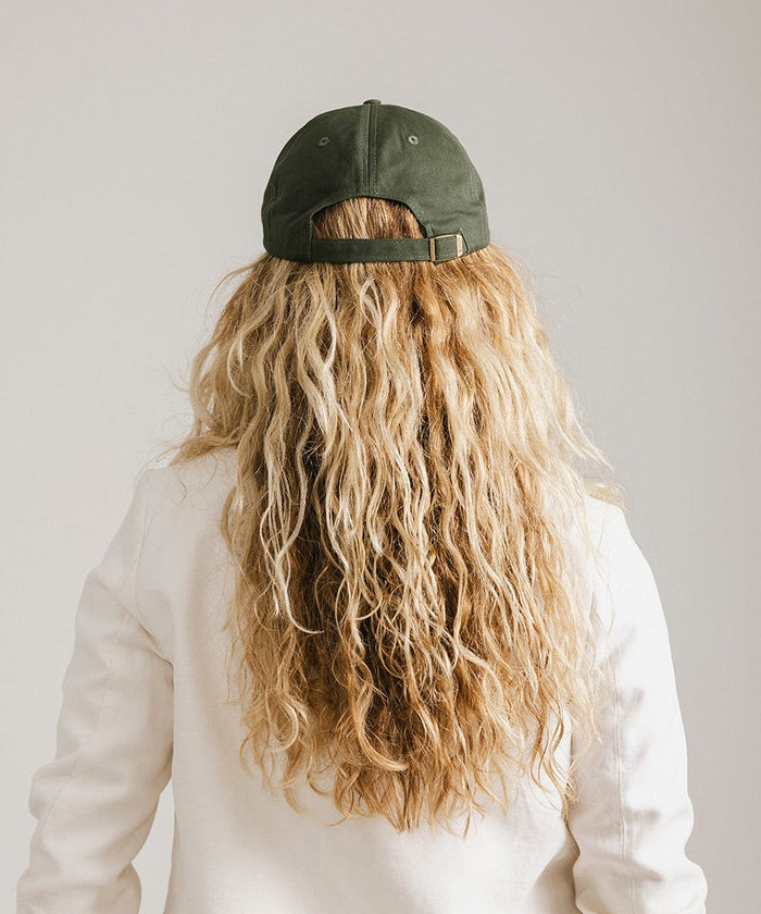 Woman wearing a dark green ball cap   and a white blazer, facing behind against a plain background  #color_dark green