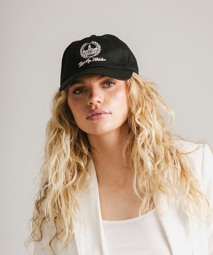 Woman wearing a black ball cap with a white logo and text and a white blazer against a plain background  #color_black
