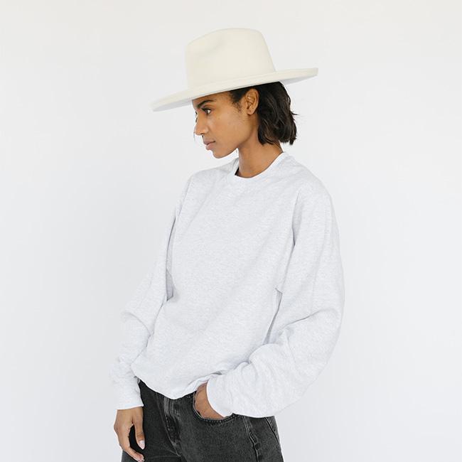 Woman wearing a white hat, an ash colored sweatshirt with the text 'GIGI PIP' in white, and dark grey jeans, facing sideways against a plain background #color_ash-white