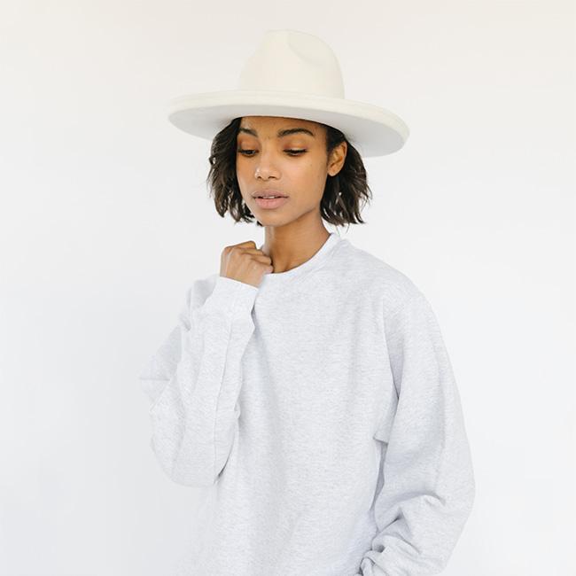 Woman wearing a white hat and an ash colored sweatshirt with the text 'GIGI PIP' in white against a plain background #color_ash-white