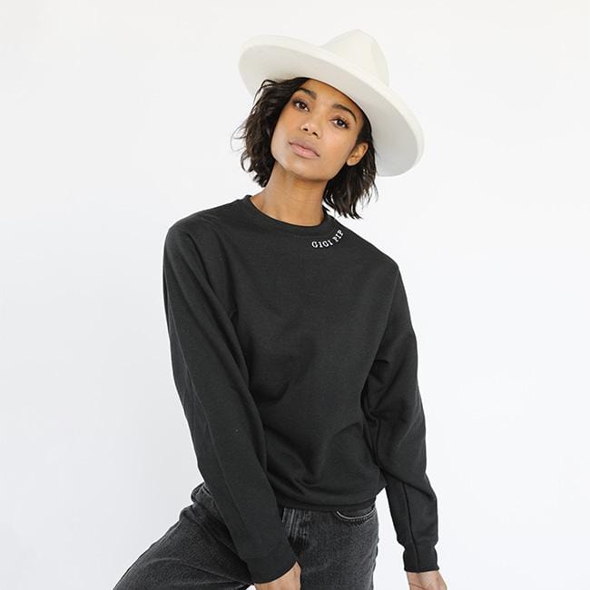 Woman wearing a white hat, a black sweatshirt with the text 'GIGI PIP' and dark grey jeans against a plain background #color_black