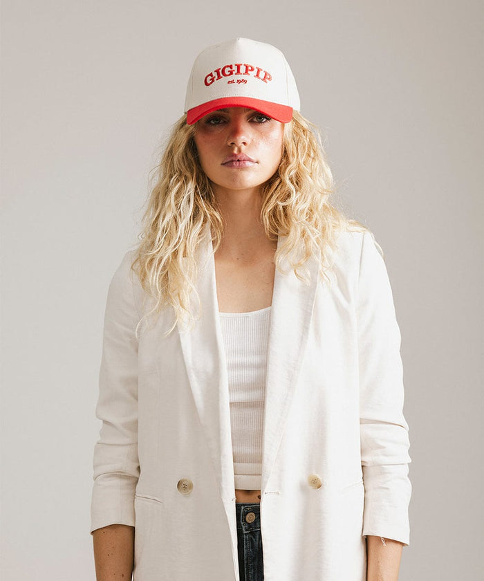 Woman wearing a cream and red trucker hat with 'GIGI PIP' embroidered in red on the front and a white coat against a plain background  #color_cream - red