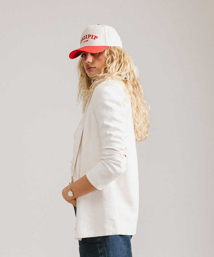 Woman wearing a cream and red trucker hat with 'GIGI PIP' embroidered in red on the front and a white coat, posing against a plain background  #color_cream - red