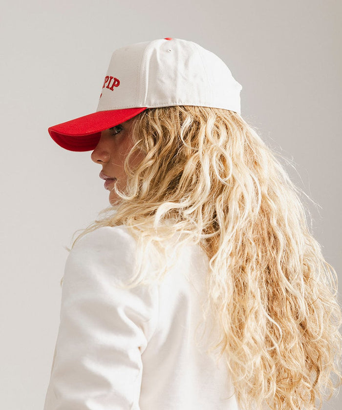 Woman wearing a cream and red trucker hat with 'GIGI PIP' embroidered in red on the front and a white coat, facing sideways against a plain background  #color_cream - red