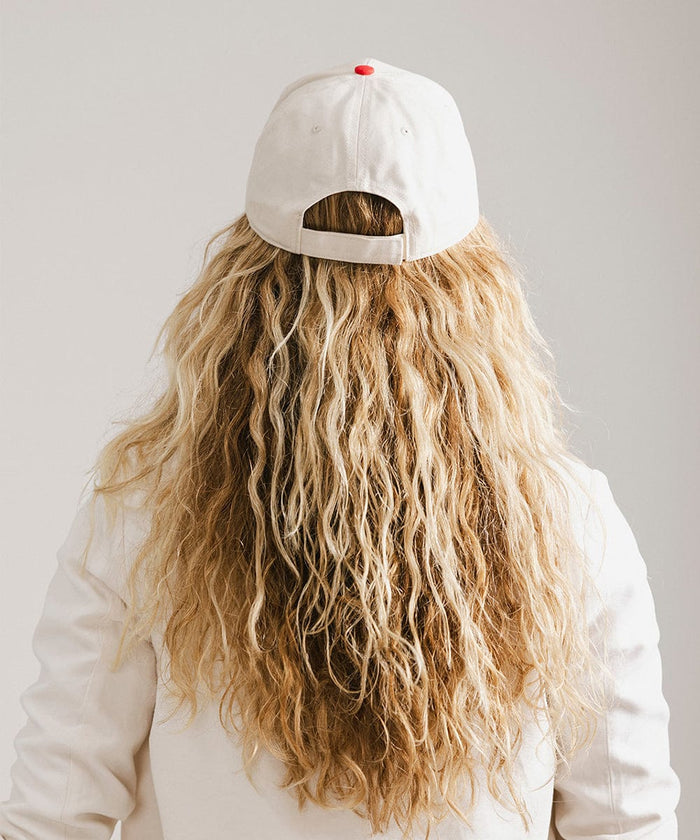 Woman with long hair wearing a cream and red trucker hat and a white coat against a plain background  #color_cream - red