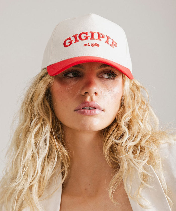 Woman wearing a cream and red trucker hat with 'GIGI PIP' embroidered in red on the front  against a plain background  #color_cream - red
