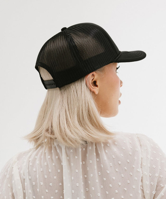 Woman wearing a black cap with golden 'GIGIRODEO' text and logo, and a white top, facing behind against a plain background #color_black