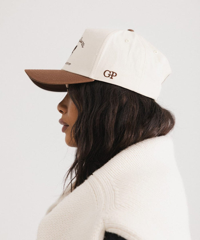 Woman wearing a cream and chocolate brown Trucker hat and a white sweater, facing sideways  against a plain background #color_cream - chocolate brown