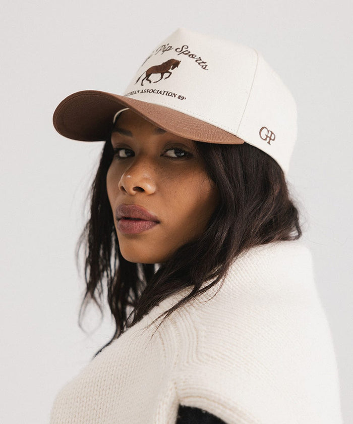 Woman wearing a cream and chocolate brown Trucker hat with 'Gigi Pip Sports' and horse logo and a denim jacket, posing sideways  against a plain background #color_cream - chocolate brown