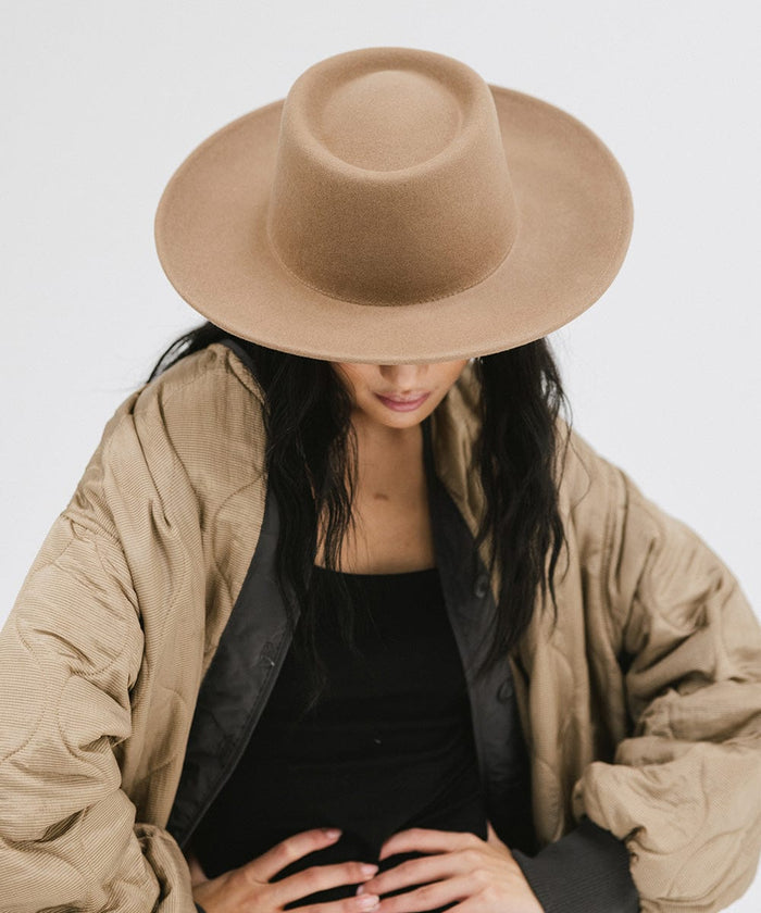 Woman wearing a brown gambler hat and a black top with a brown jacket, looking down against a plain background #color_brown