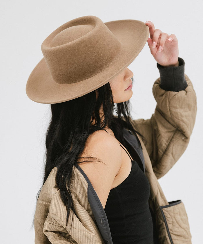 Woman wearing a brown gambler hat and a black top with a brown jacket, facing sideways against a plain background #color_brown